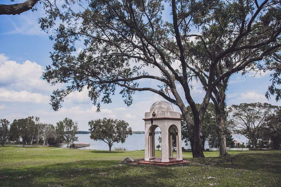 A peaceful outdoor scene at Lake Gibson Village featuring a small white gazebo with arches and a dome roof, surrounded by large trees and green grass, with a lake and a dock visible in the background under a partly cloudy sky.