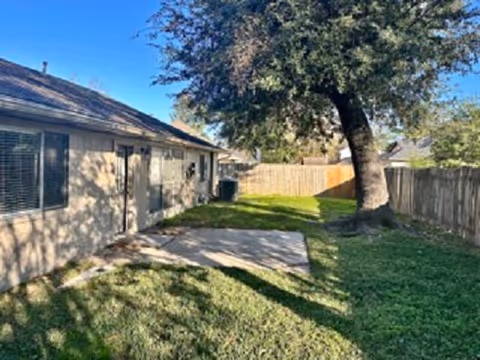 Backyard of a single-story house with a grassy lawn, a small concrete patio, a large tree, and a wooden fence.
