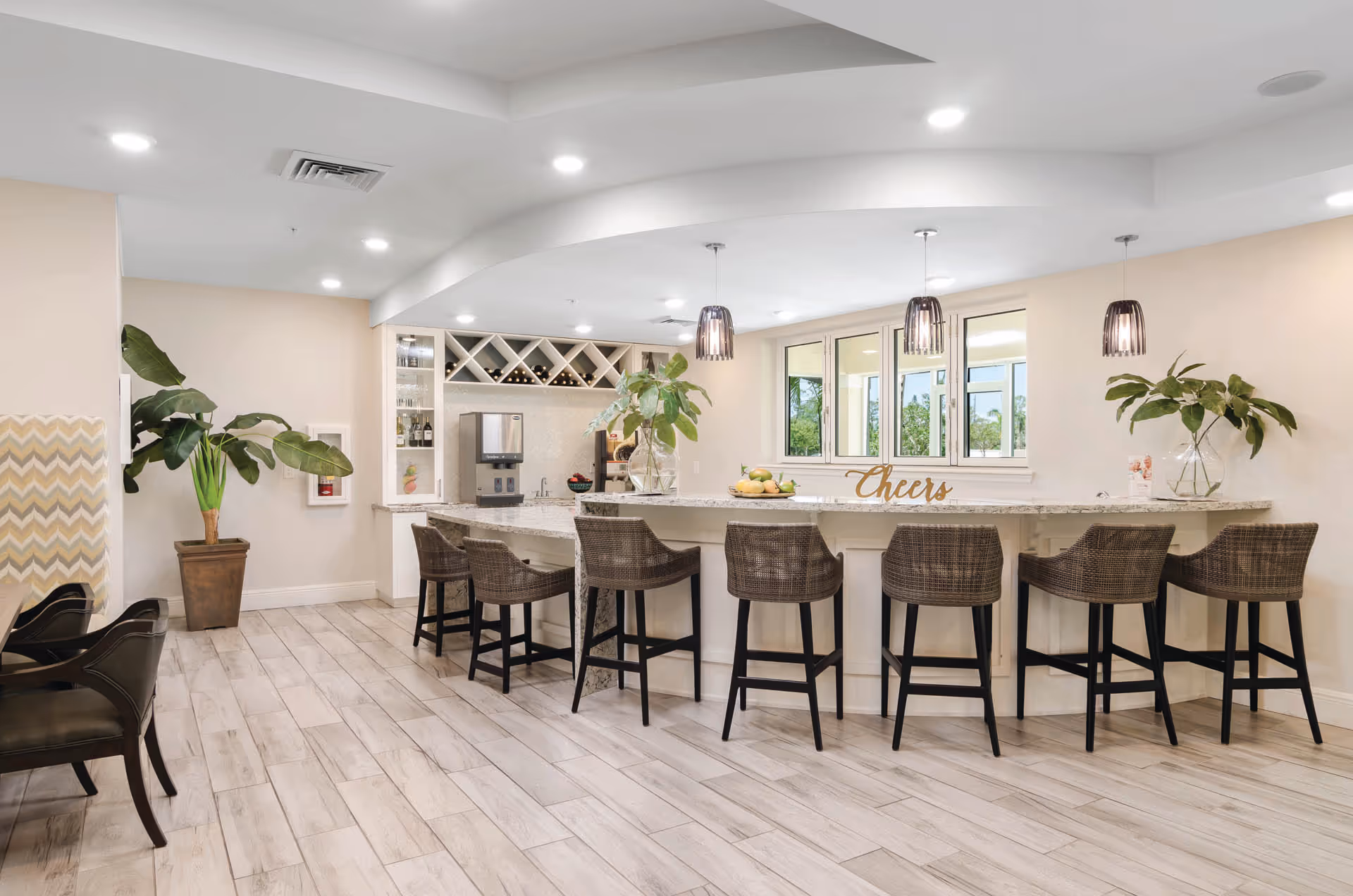 A modern, bright bar area with a long marble countertop and seven wicker bar stools. The back wall features a wine rack with bottles, a beverage dispenser, and glass shelves. Two large plants in glass vases and a decorative 'Cheers' sign are placed on the counter. The floor is light wood tile, and the ceiling has recessed lighting and three pendant lights hanging above the bar. Large windows provide a view of greenery outside.