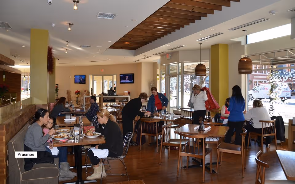 Interior of a dining room with several people eating and socializing at wooden tables and chairs. Large windows let in natural light, and there are pendant lights hanging from the ceiling. The room has a warm and inviting atmosphere with wooden flooring and a decorative wooden ceiling feature.