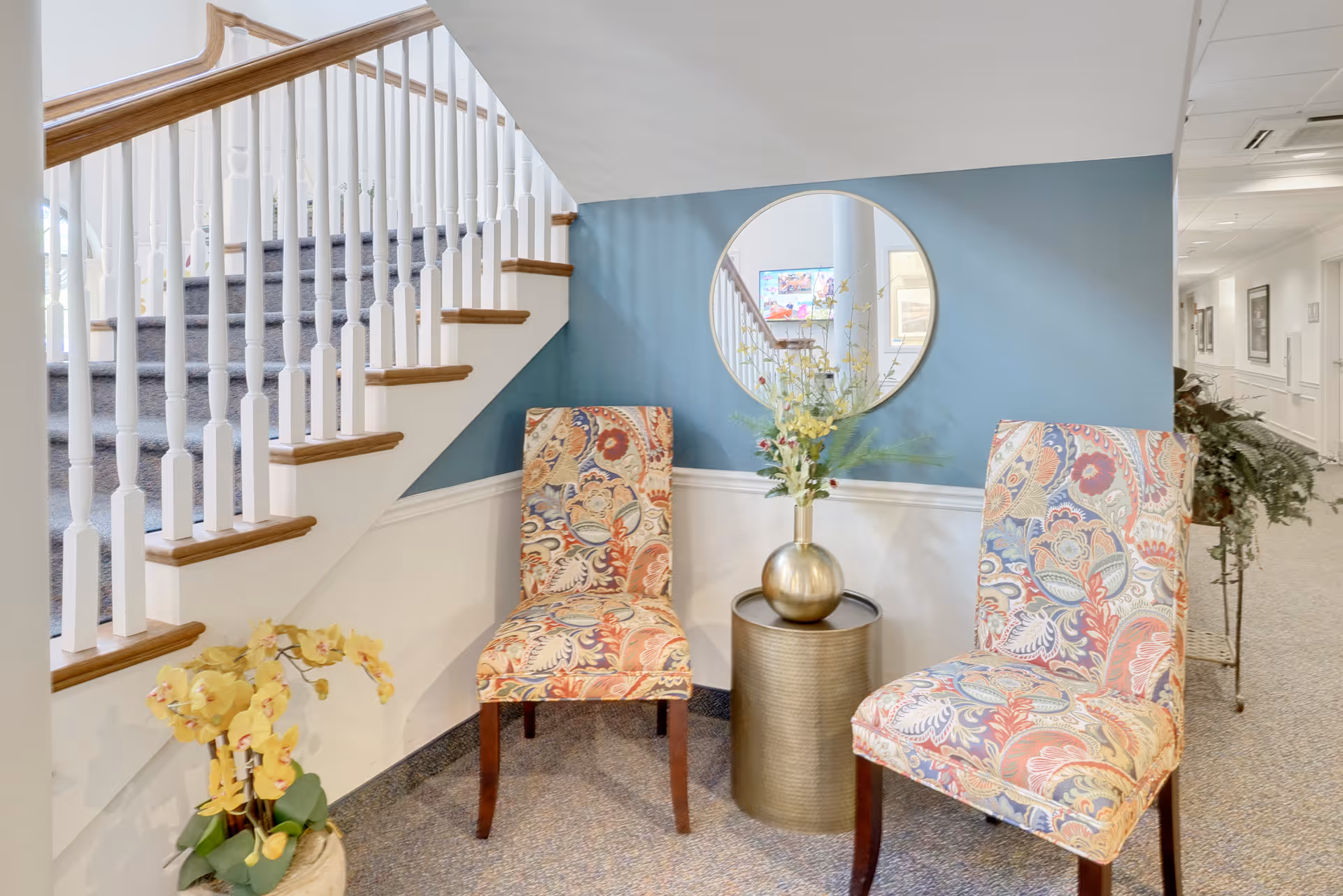 A cozy seating area under a staircase with two patterned upholstered chairs and a round metallic side table holding a vase with flowers. A round mirror hangs on the blue accent wall behind the chairs, reflecting part of the staircase and a hallway with framed pictures.