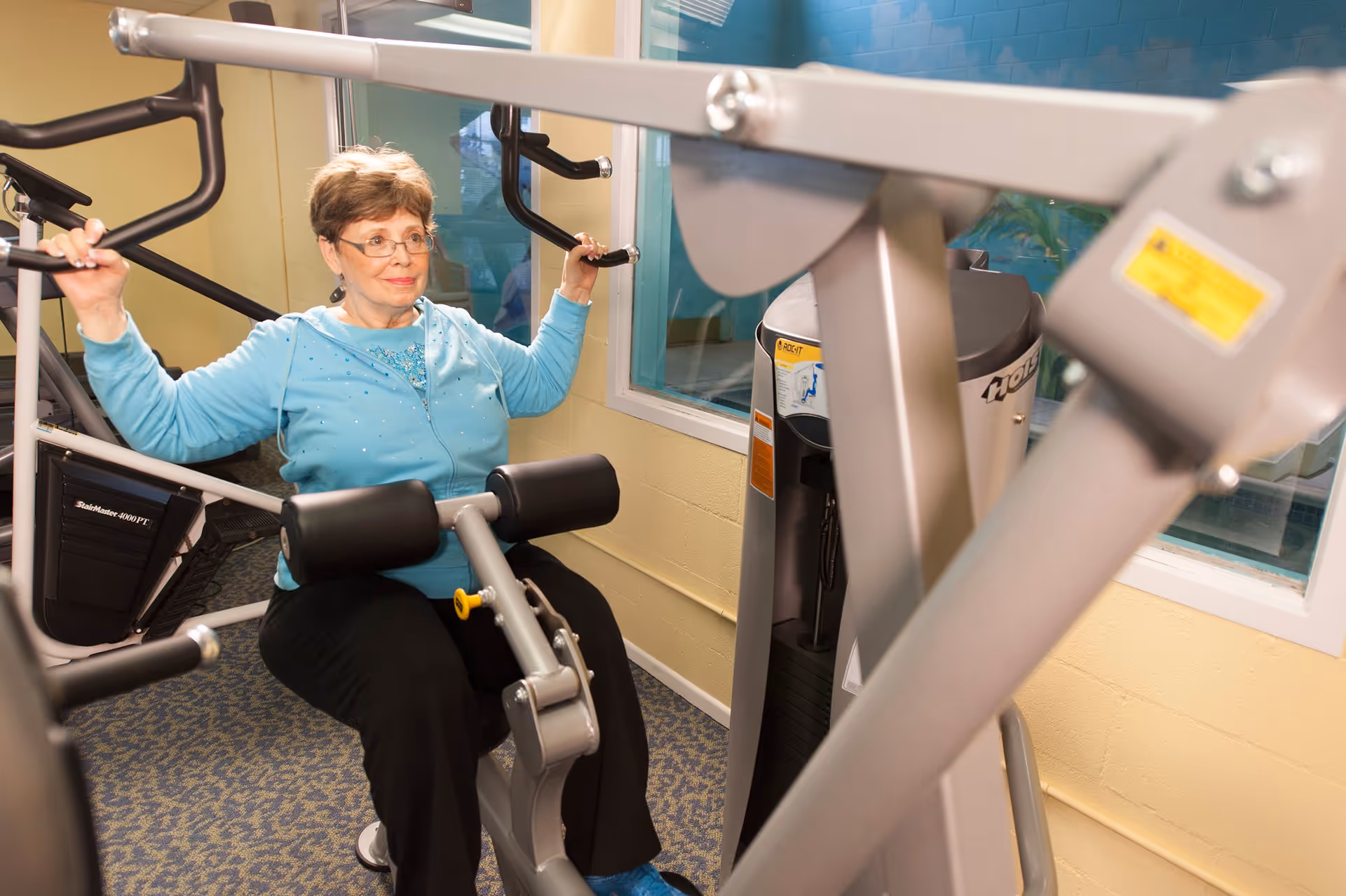 An elderly woman wearing glasses and a light blue hoodie is using a strength training machine in a fitness room with carpeted floors and light yellow walls. There is a window behind her and other exercise equipment visible in the background.