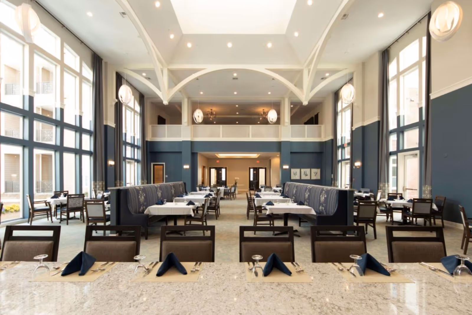A spacious and elegant dining room in a senior living facility with high ceilings, large windows allowing natural light, and modern pendant lights. The room features tables set with white tablecloths, navy blue napkins, and silverware, along with cushioned chairs and booth seating with floral upholstery.