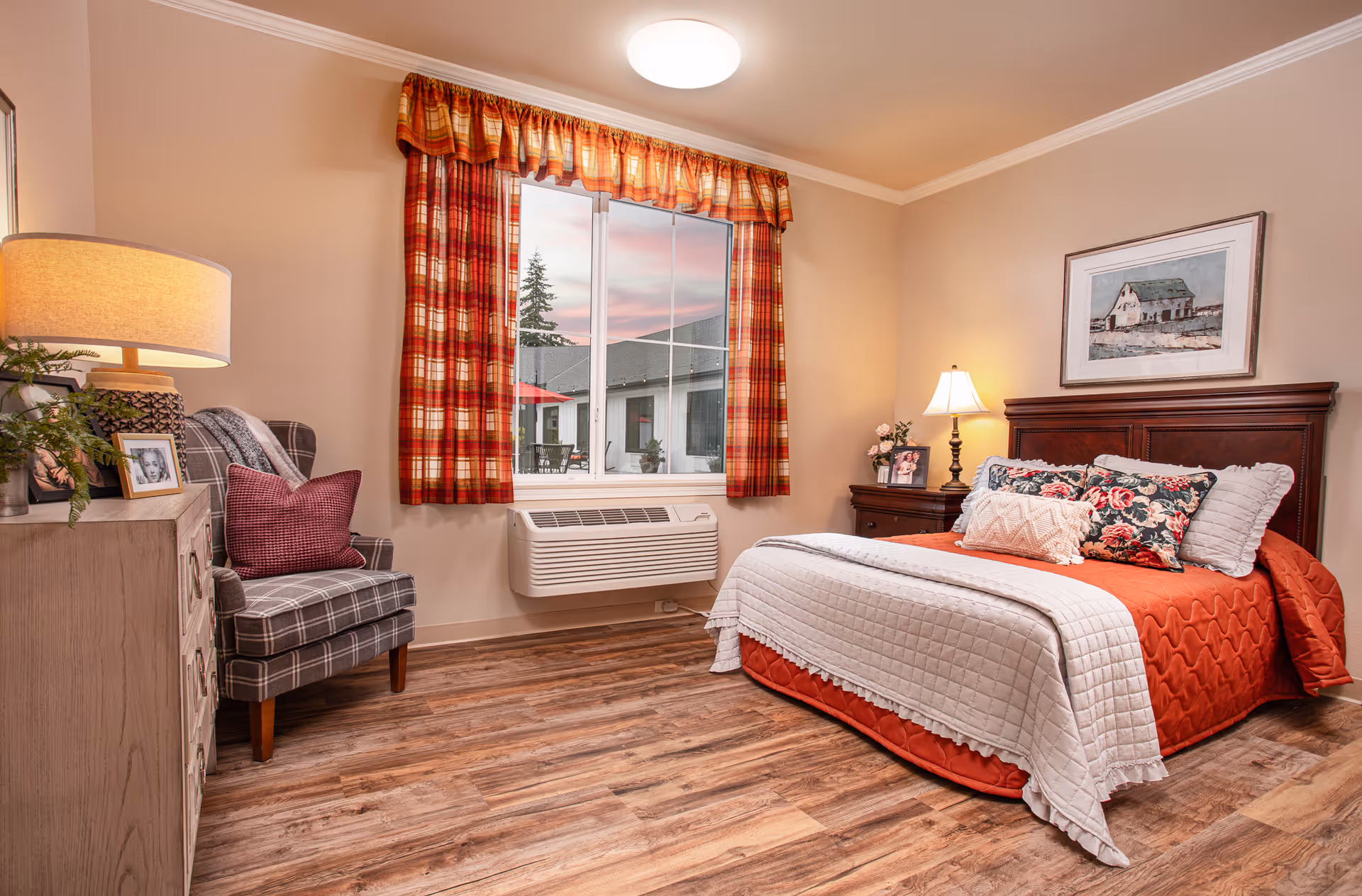 A cozy bedroom with a wooden bed frame, orange and white bedding, floral pillows, a nightstand with a lamp and framed photo, a plaid armchair with a red pillow, a dresser with a lamp and framed photos, and a window with red plaid curtains showing an outdoor patio area.