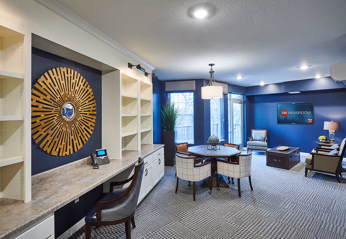 A bright common living area with a round table and chairs, built-in desk and shelving, lounge seating, and a wall-mounted TV.