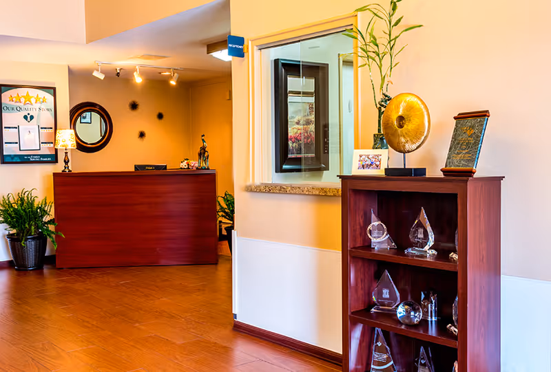 Reception area with a wooden front desk, a round mirror on the wall, a lamp, and plants. To the right, a wooden shelf displays various glass awards and decorative items, with a framed photo and a plant on top. The walls are painted in warm tones and the floor has wood-like flooring.