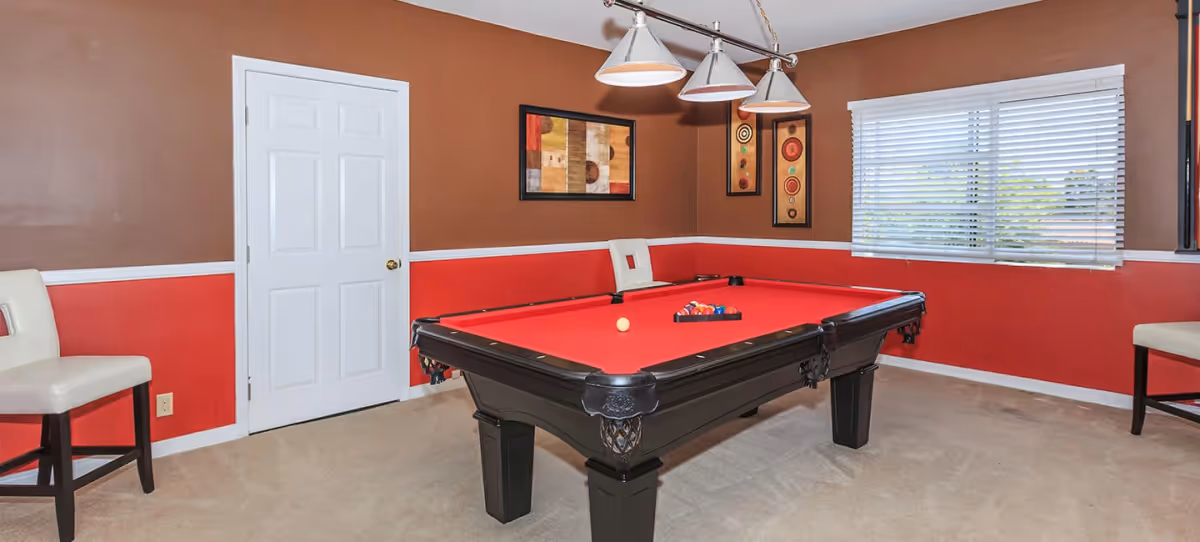 A game room with a red felt pool table in the center, surrounded by three white cushioned chairs. The walls are painted brown on the upper half and red on the lower half, with white trim. There are three pendant lights hanging above the pool table and three framed artworks on the walls. A window with white blinds is on the right side, allowing natural light into the room.