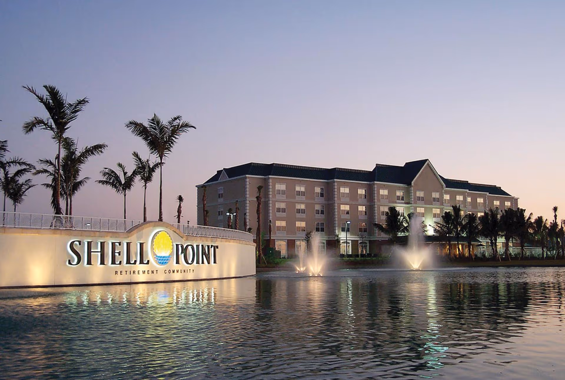 Exterior view of the Shell Point Welcome Center at dusk with an illuminated entrance sign, palm trees, and fountains reflecting in a pond.