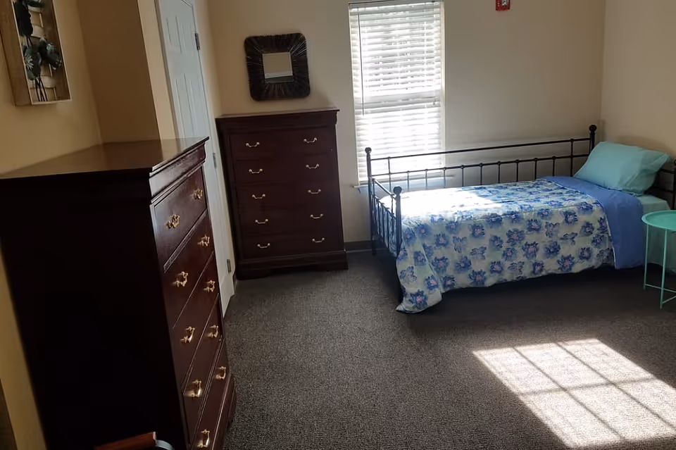 Sunlit bedroom with a single metal-framed bed covered in a floral bedspread, two dark wood dressers, and a window.
