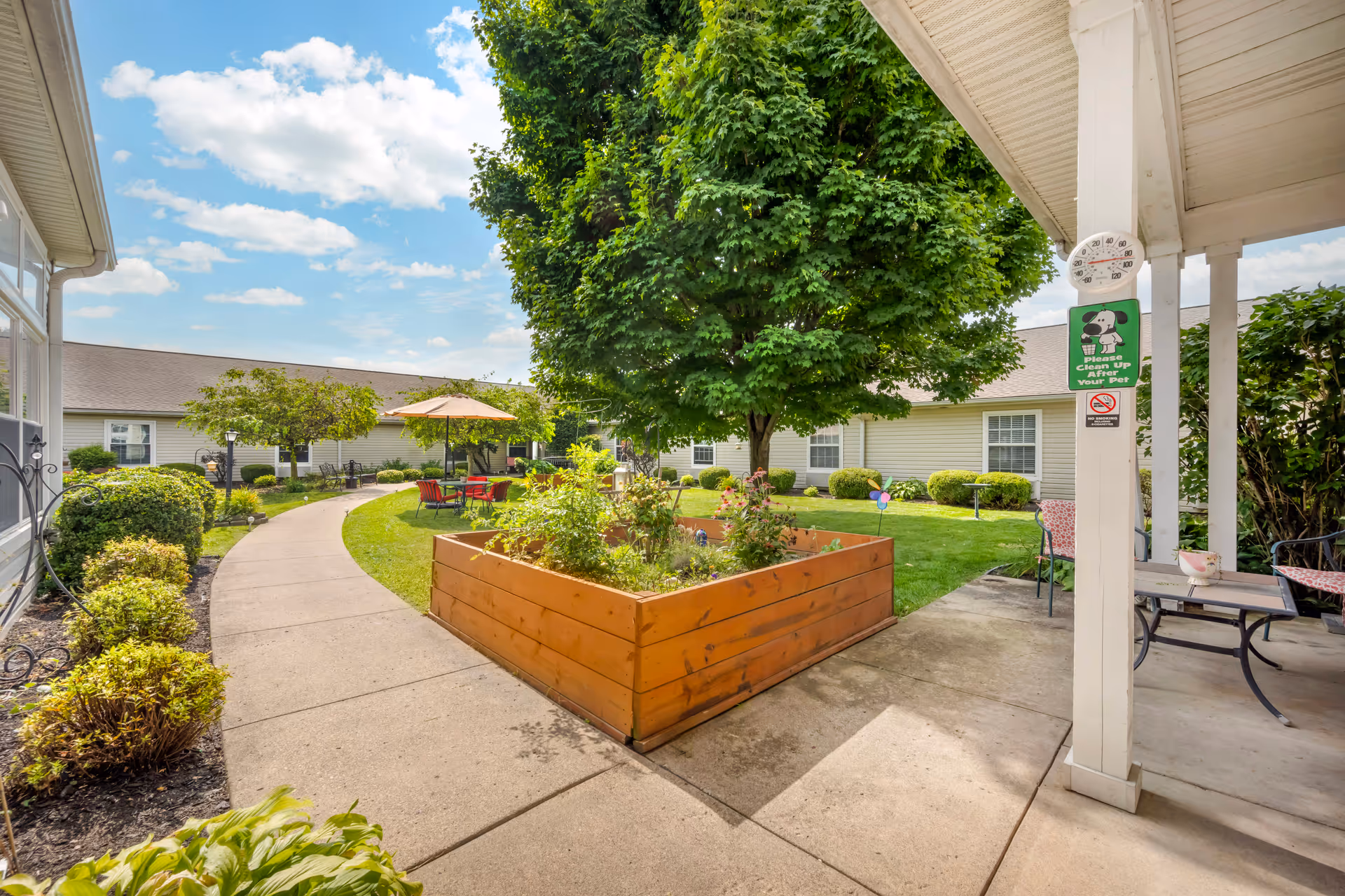Outdoor courtyard area at Brookdale Buck Creek featuring a large green tree, a raised wooden garden bed with plants, a curved concrete walkway, patio tables with umbrellas and chairs, and well-maintained landscaping under a partly cloudy sky.