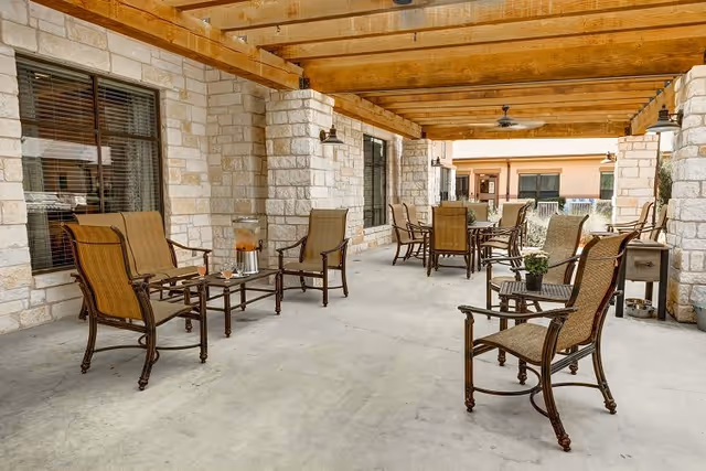 Covered outdoor patio area with stone pillars and wooden beams overhead, furnished with multiple sets of brown metal and wicker chairs and tables. There is a beverage dispenser on one table and a small potted plant on another. The area has concrete flooring and windows along the stone wall.