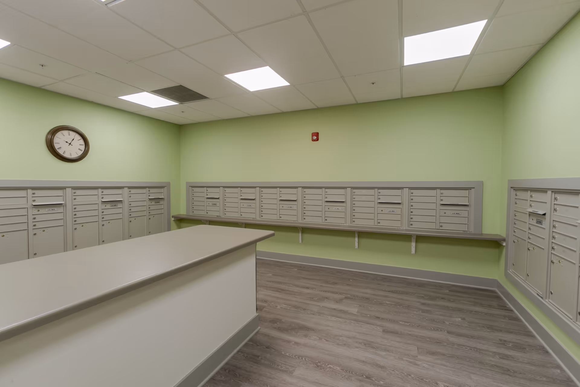 A mailroom with light green walls and gray mailboxes mounted on three walls. There is a large gray counter in the center of the room and a round wall clock above the mailboxes on the left wall. The floor has wood-like vinyl flooring and the ceiling has recessed lighting panels.