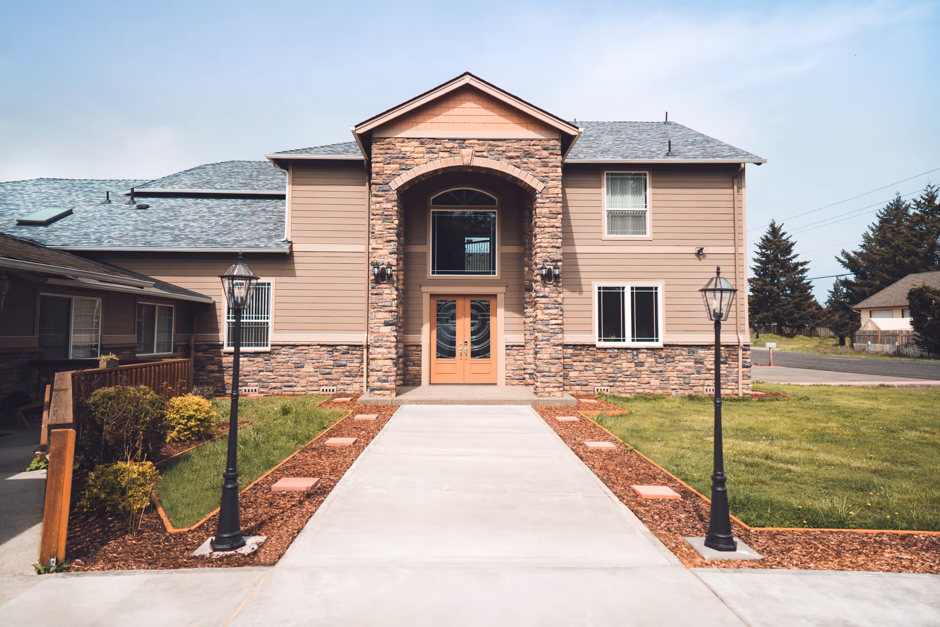 Front exterior of a two-story residential building with a stone entryway, double doors and a concrete walkway flanked by lamp posts.