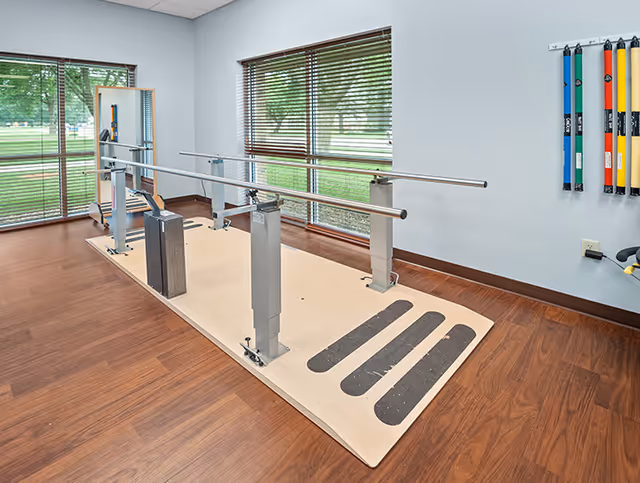 A physical therapy room with parallel walking bars on a mat and large windows overlooking a lawn.