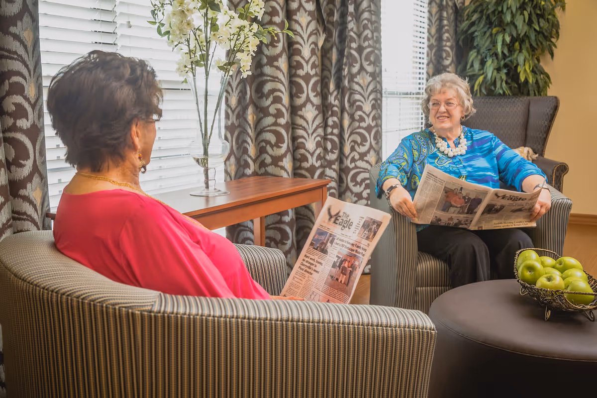 Two elderly women sitting in a cozy living room area, each holding a newspaper. One woman is wearing a bright pink top and the other is wearing a blue patterned blouse with a white necklace. They are seated in striped armchairs near a window with patterned curtains. A round table with a decorative bowl of green apples is in front of them, and a wooden side table with a vase of white flowers is behind them.