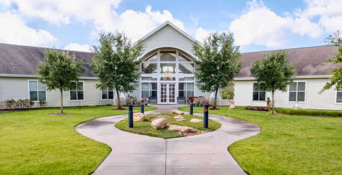 Front entrance of a single-story assisted living building with a curved walkway, lawn, small trees, and a glass entryway.