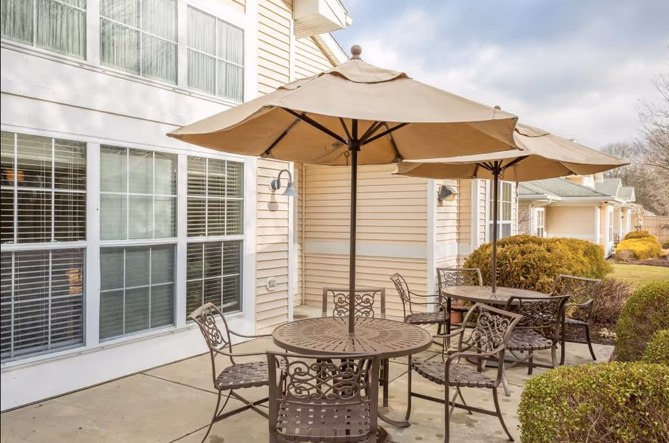 Outdoor patio area with two round metal tables, each surrounded by four metal chairs with decorative backs. Each table has a large beige umbrella providing shade. The patio is adjacent to a beige building with large windows and white trim. There are bushes and greenery around the patio area under a partly cloudy sky.