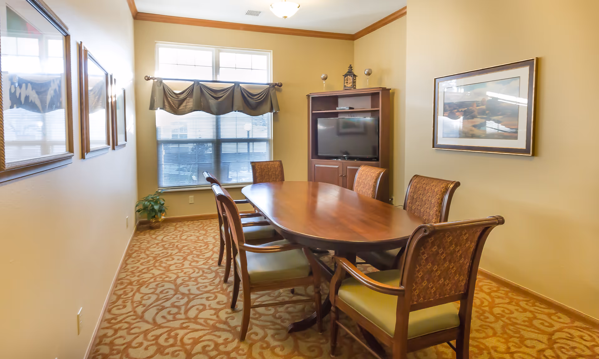 A dining room with a long wooden table surrounded by six upholstered chairs. The room has beige walls, a patterned carpet, a window with blinds and a valance, a wooden cabinet with a TV on top, and framed artwork on the walls.