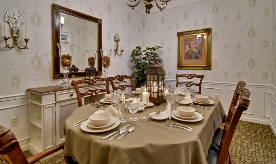 A dining room with a table set for six people, featuring plates, bowls, glasses, silverware, and folded napkins. The table is covered with a beige tablecloth and has a decorative lantern and candles in the center. The room has patterned wallpaper, a large mirror on one wall, a framed floral painting, and a sideboard with decorative glass vases filled with potpourri. There is also a green potted plant in the corner.