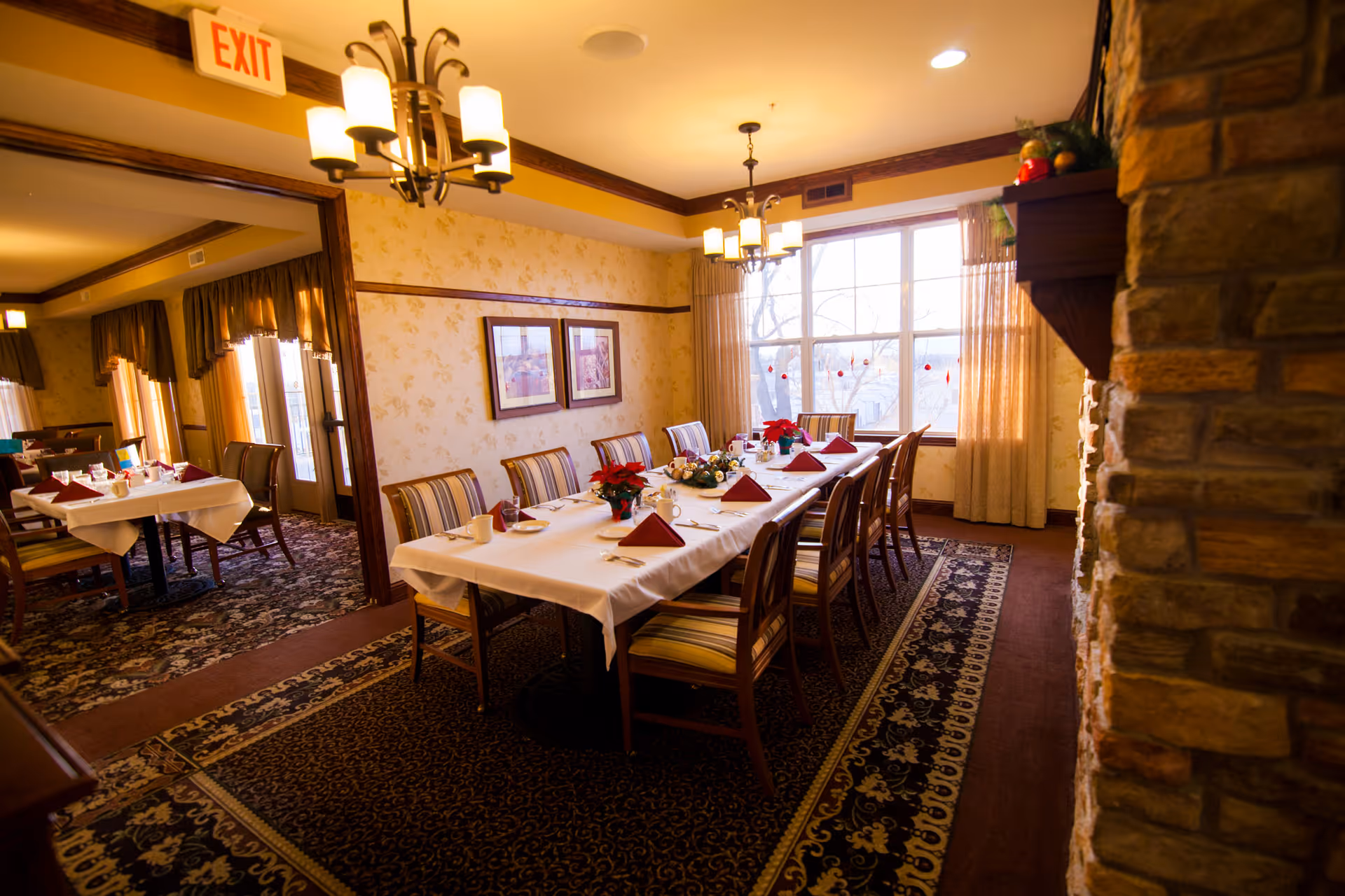 A warmly lit dining room with a long table set for a meal, featuring white tablecloths, red folded napkins, cups, and floral centerpieces. The room has patterned carpet, striped upholstered chairs, framed pictures on the wall, large windows with curtains, and a stone fireplace on the right side. Another dining area is visible through an open doorway.