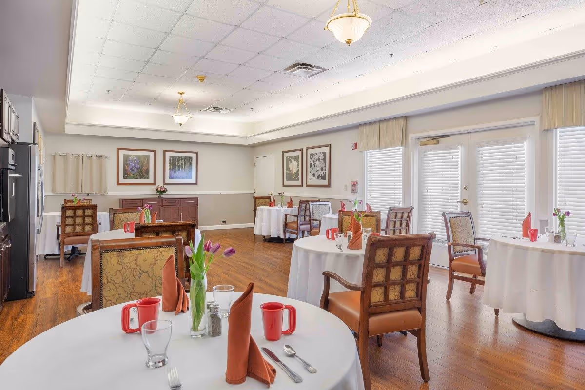 A bright dining room with round tables covered in white tablecloths, each set with red mugs, glasses, silverware, and folded brown napkins. The room has wooden floors, several wooden chairs with patterned upholstery, framed floral artwork on the walls, and large windows with blinds letting in natural light. There are also fresh flowers in vases on the tables.