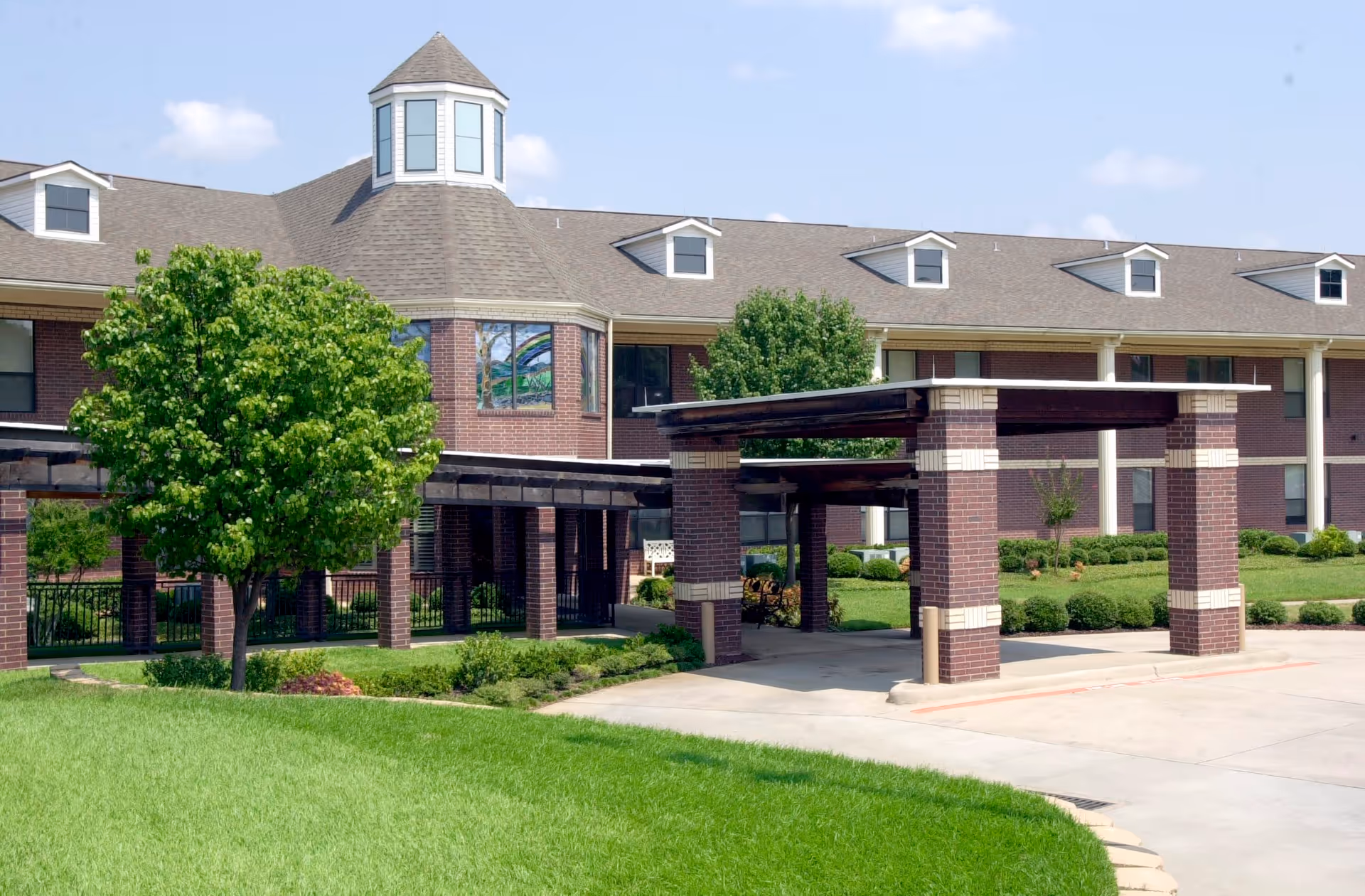Exterior view of Buckner Westminster Place, a brick building with a covered entrance supported by brick columns, surrounded by green grass, trees, and shrubs under a partly cloudy sky.