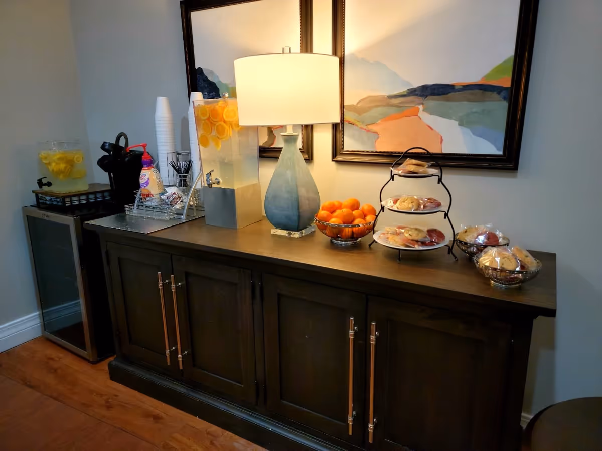 A wooden sideboard with a blue table lamp, a large beverage dispenser filled with lemon water, a bowl of oranges, and several bowls and a tiered tray with individually wrapped cookies. Behind the sideboard are two framed abstract landscape paintings. To the left, there is a small refrigerator and another beverage dispenser with lemon water.