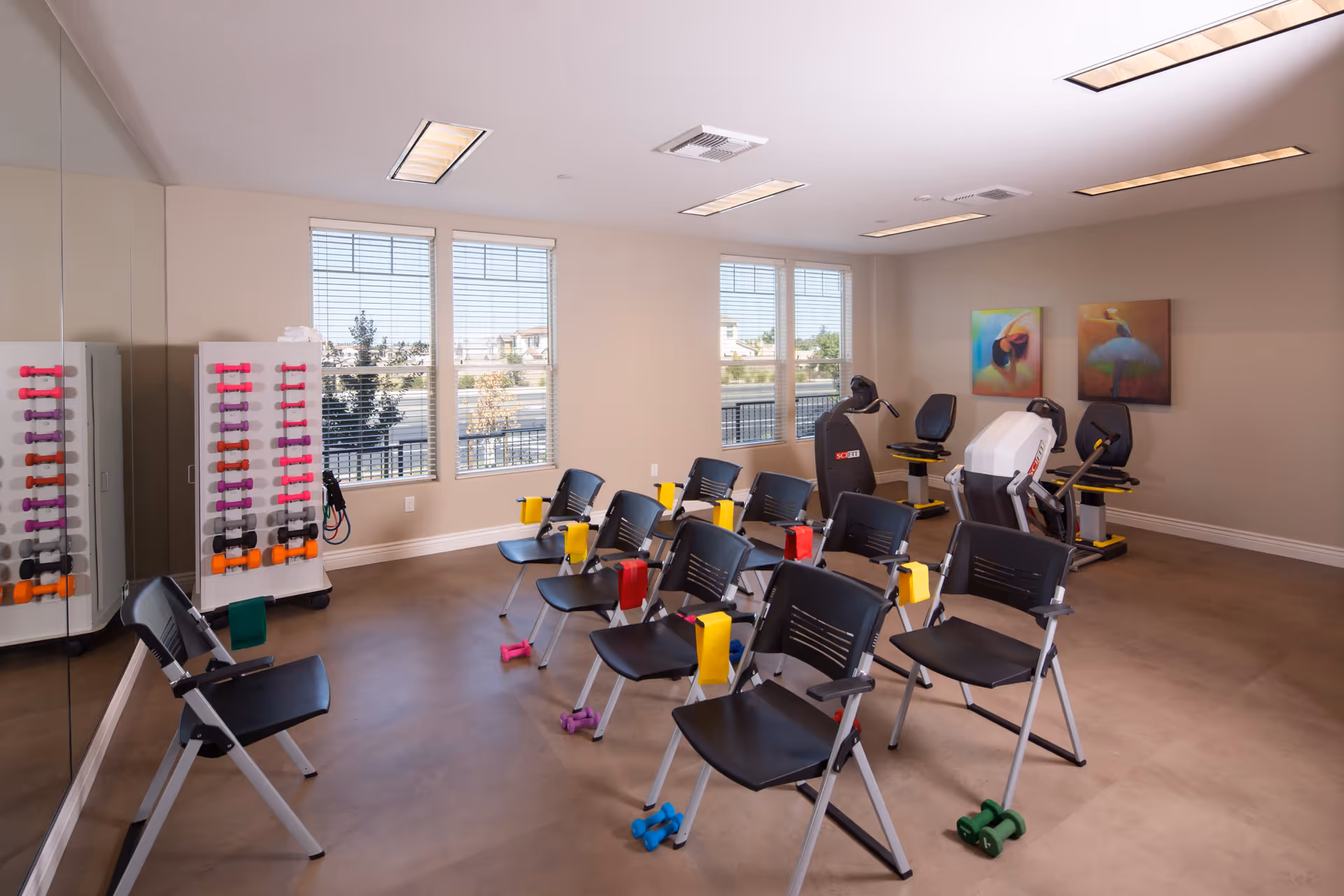 A bright exercise room with several black chairs arranged in rows, each paired with colorful dumbbells on the floor. The room has large windows letting in natural light, a rack of various colored dumbbells against the wall, and two exercise machines in the corner. Two colorful paintings hang on the far wall.