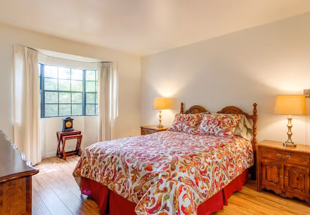 A cozy bedroom with a wooden bed frame and a colorful patterned bedspread. There are two wooden nightstands on either side of the bed, each with a lamp. A large window with white curtains lets in natural light, and a small wooden table with a decorative clock is placed in front of the window. The room has wooden flooring and light-colored walls.