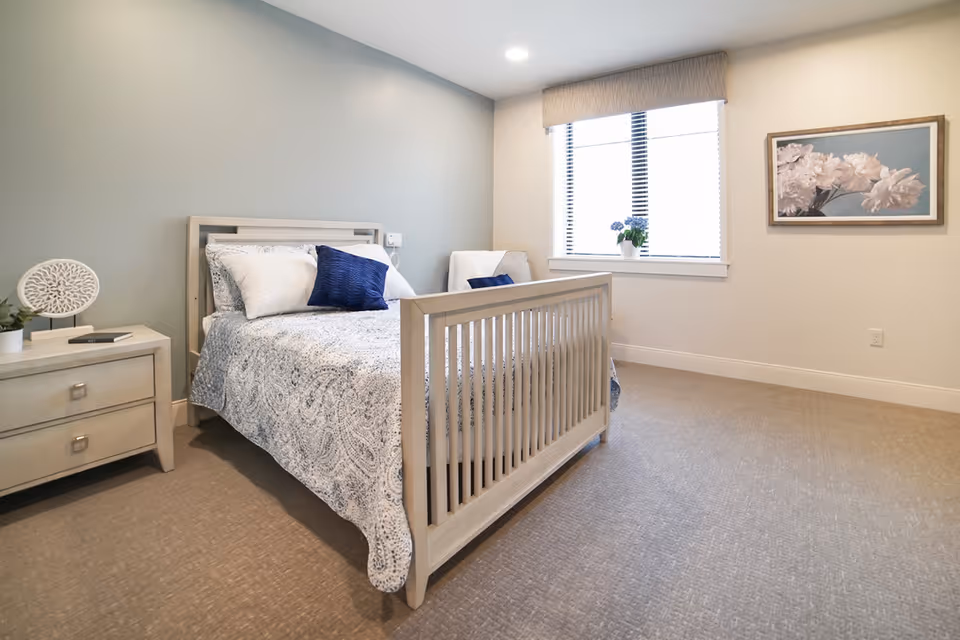 A neatly made bed with white and blue patterned bedding and pillows in a bright bedroom. There is a light-colored wooden nightstand with a decorative item and a book, a window with blinds and a potted plant on the sill, a white armchair with a blue pillow, and a framed floral picture on the wall.