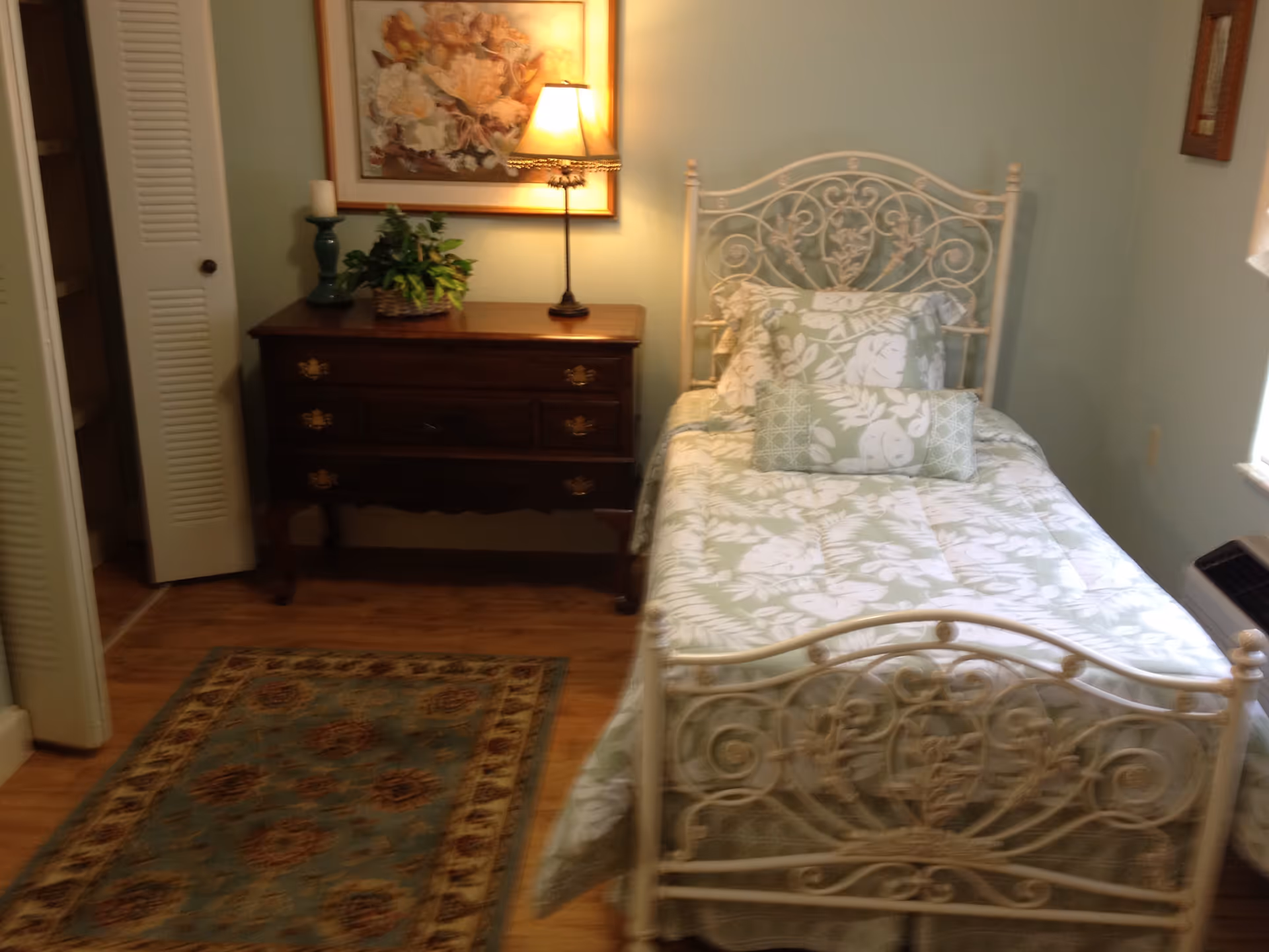 Small bedroom with a single ornate metal bed dressed in floral bedding, a wooden dresser with a lamp and plant, and a patterned rug on hardwood floor.