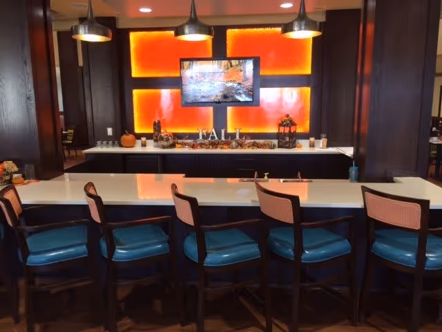 Interior view of a modern dining or common area with a long white countertop and six high chairs with teal seats and pink backs. Behind the counter is a dark wood cabinet with a bright orange illuminated panel and a mounted flat-screen TV. The counter is decorated with fall-themed items including a pumpkin, lantern, and the word 'FALL' spelled out in letters.