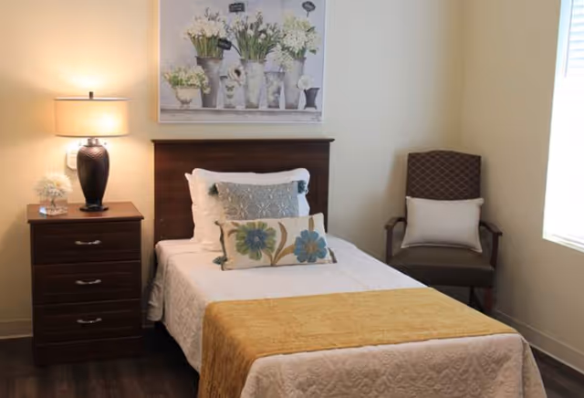 A neatly made single bed with white bedding, decorative pillows, and a yellow throw blanket in a cozy room. To the left of the bed is a wooden nightstand with a lamp and a small flower arrangement. Above the bed hangs a framed picture of white flowers in vases. To the right of the bed is a cushioned armchair with a white pillow, next to a window with blinds.