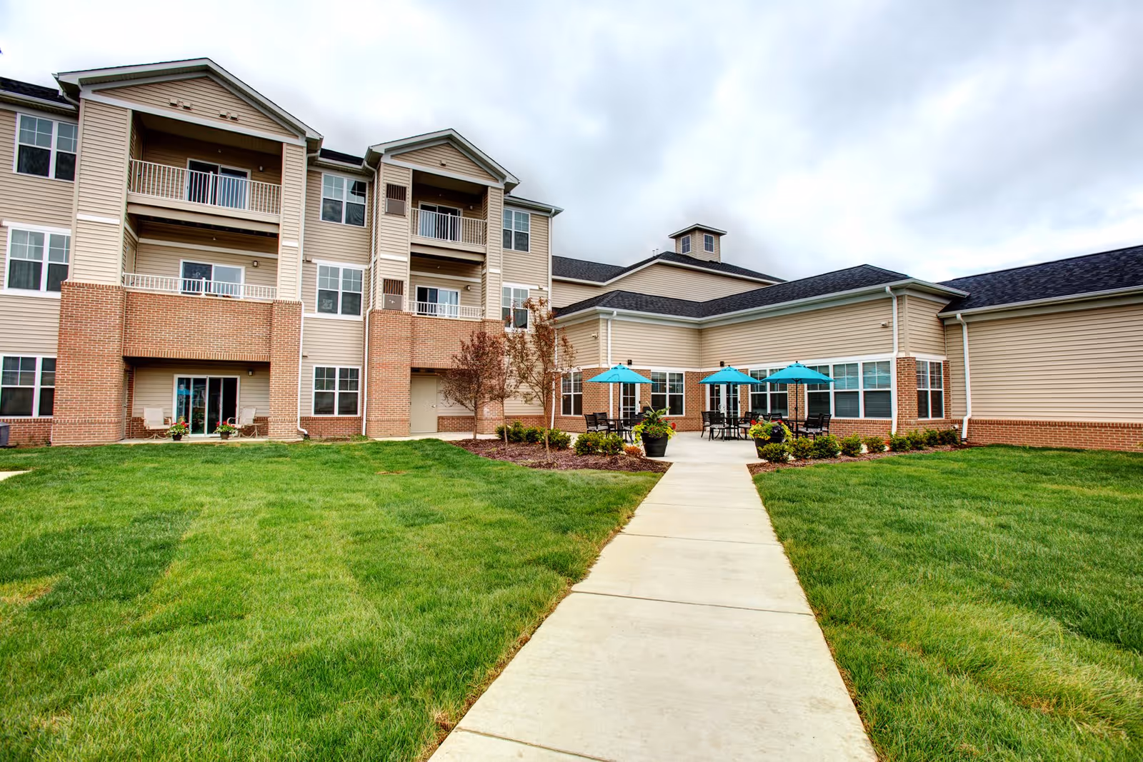 Exterior view of a multi-story senior living facility with beige siding and brick accents. There is a concrete walkway leading to a patio area with tables and turquoise umbrellas. The surrounding area has well-maintained green grass and some small trees and shrubs under a cloudy sky.