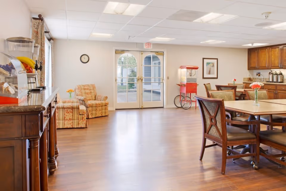 A bright and spacious common area with wooden flooring, featuring a seating area with two patterned armchairs near a window, a dining area with tables and chairs, a popcorn machine, and a wooden cabinet with a water dispenser and fruit on top. Double glass doors lead outside.