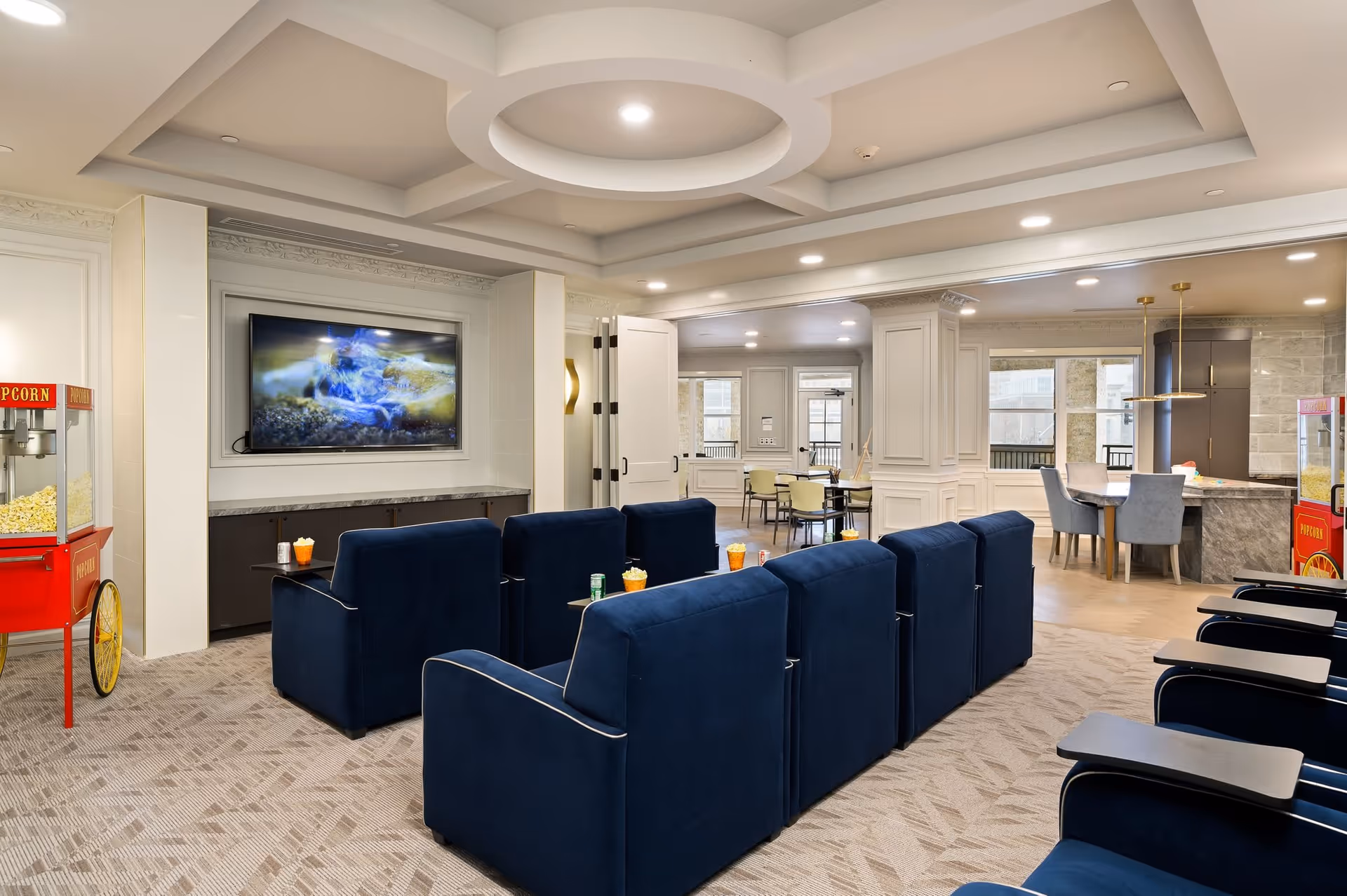 A modern common area with rows of blue upholstered theater-style chairs facing a wall-mounted flat screen TV. There are popcorn machines on either side of the room. The space has a coffered ceiling with recessed lighting and an open layout leading to a dining area with tables and chairs.