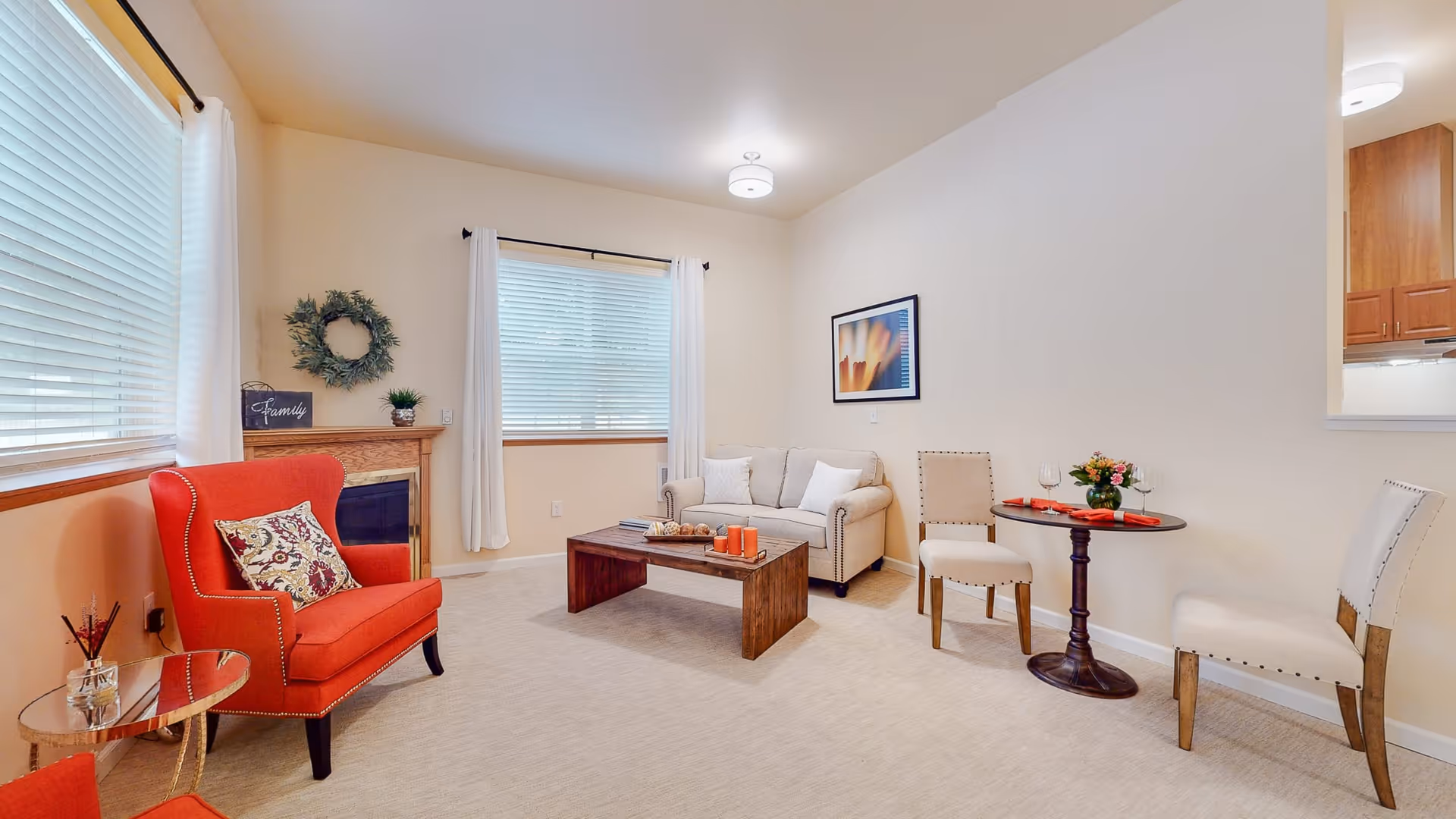 A cozy living room with a red armchair, a beige loveseat, a wooden coffee table, and a small round table with two beige chairs. The room has two windows with white curtains, a decorative wreath on the wall, and a framed abstract artwork. The walls are light-colored, and the floor is carpeted.