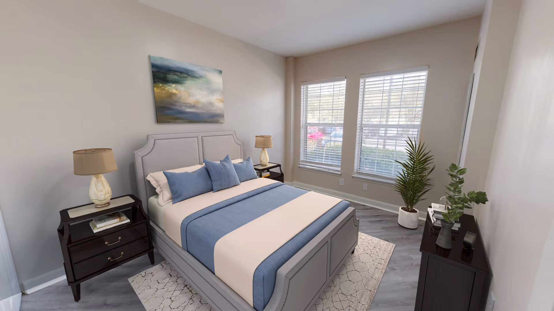 A neatly arranged bedroom with a gray upholstered bed featuring blue and beige bedding. There are two dark wooden nightstands on either side of the bed, each with a lamp. A large abstract painting hangs above the bed. The room has two large windows with blinds, a light-colored rug under the bed, and two potted plants near a dark wooden dresser.