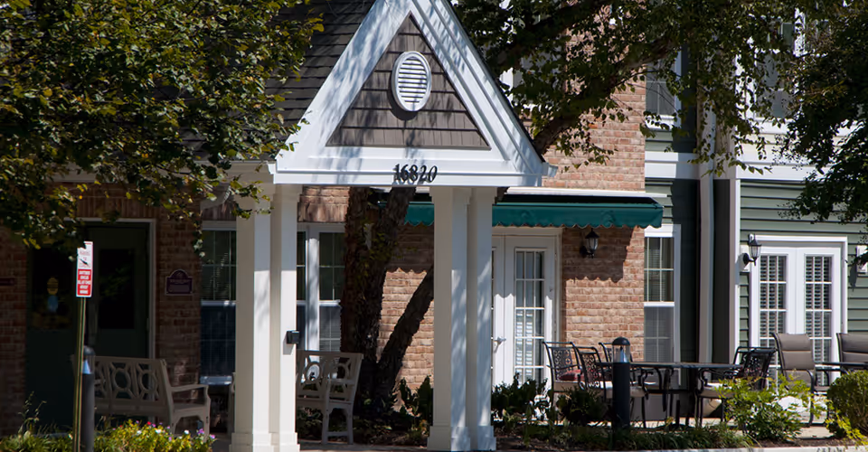 Front entrance of a senior living building with a white portico, green awnings, outdoor seating and surrounding trees.