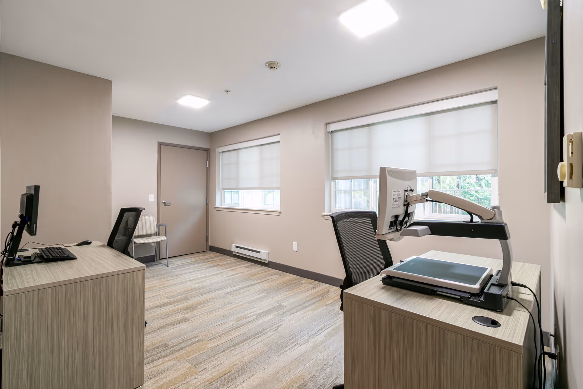 Small office room with two desks, computer monitors, chairs, windows with blinds, and a closed door.