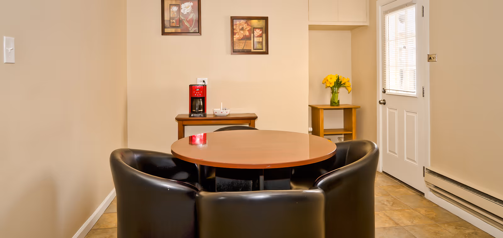 A small dining area with a round wooden table surrounded by four black leather chairs. In the background, there is a small wooden table with a red coffee maker and a bowl on it. On the right side, there is a wooden side table with a vase of yellow flowers and a white door with a window. Two framed floral pictures hang on the beige wall above the coffee maker.