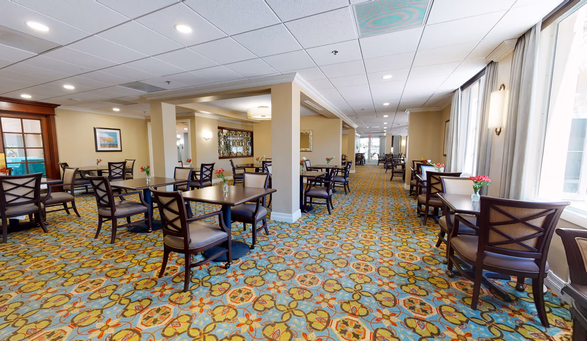 A spacious dining room with multiple dark wood tables and chairs arranged neatly on a colorful patterned carpet. The room has beige walls, large windows with curtains on the right side, and ceiling lights providing bright illumination. Small flower vases are placed on each table, and framed artwork and mirrors decorate the walls.