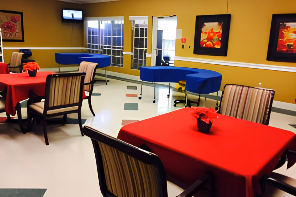 Interior view of a common dining area with tables covered in red tablecloths and striped cushioned chairs. The room has yellow walls adorned with framed artwork, a wall-mounted TV in the corner, and large windows with white trim. There is a blue curved table or counter along one wall and a mirror reflecting part of the room.
