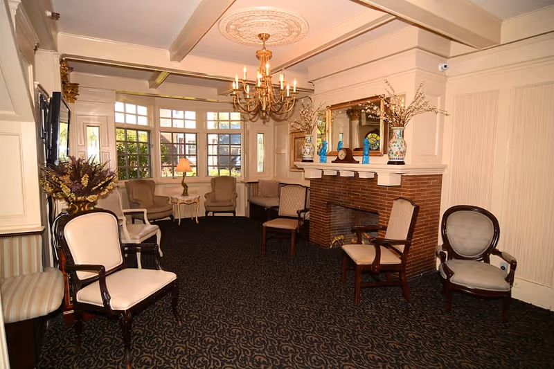 A cozy living room area with multiple upholstered chairs arranged around a brick fireplace. The room features a large window letting in natural light, a chandelier hanging from an ornate ceiling medallion, a large mirror above the fireplace, and decorative vases with flowers on the mantel. The carpet has a dark patterned design, and the walls are painted in a light cream color.