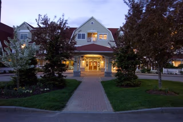 Front exterior view of a two-story senior living facility building at dusk with a covered entrance, illuminated windows, a brick walkway, and landscaped greenery including trees and shrubs.