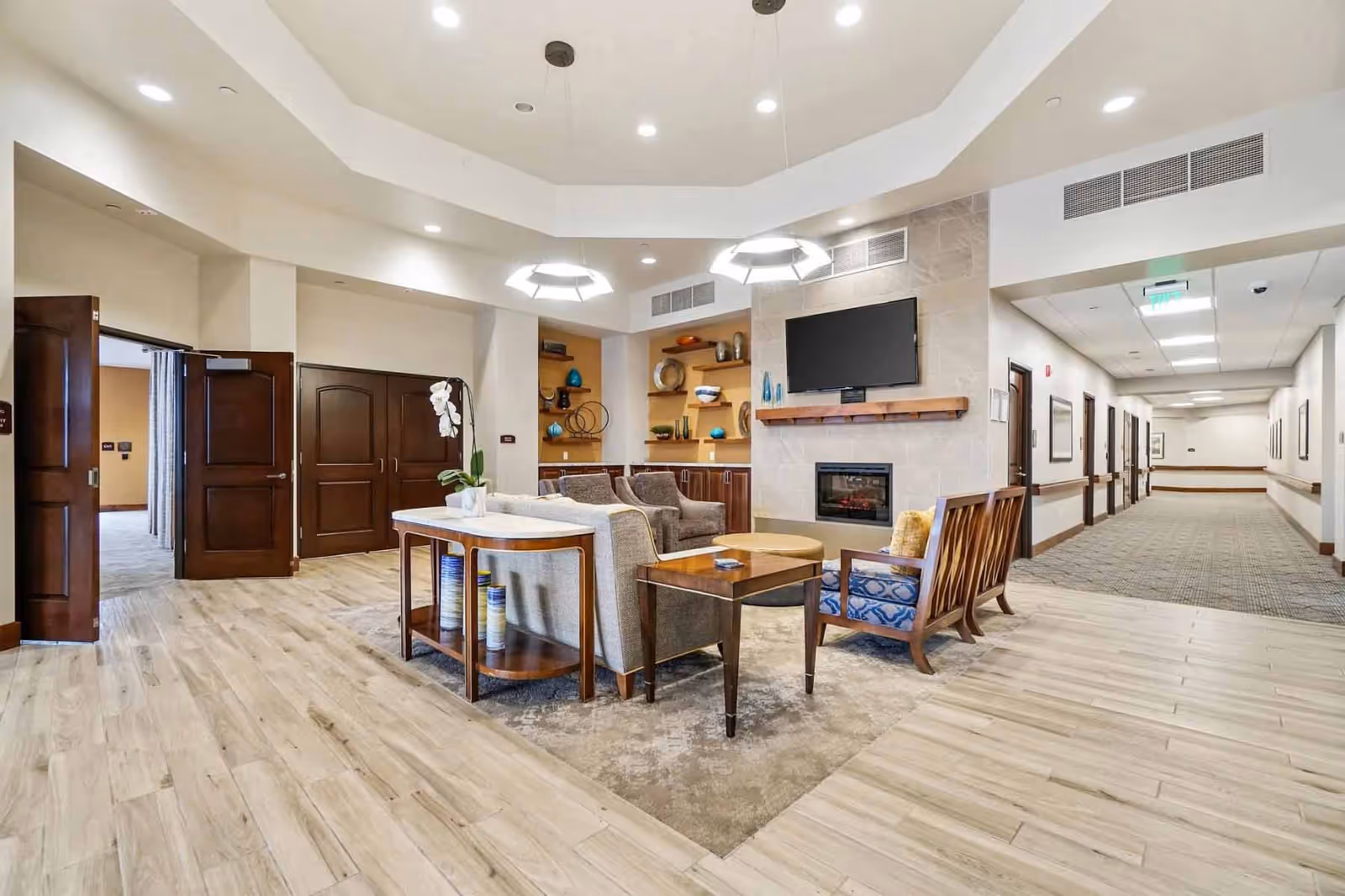 Bright communal lounge with sofas and chairs facing a TV mounted above a fireplace, shelving, and a hallway leading to resident rooms.