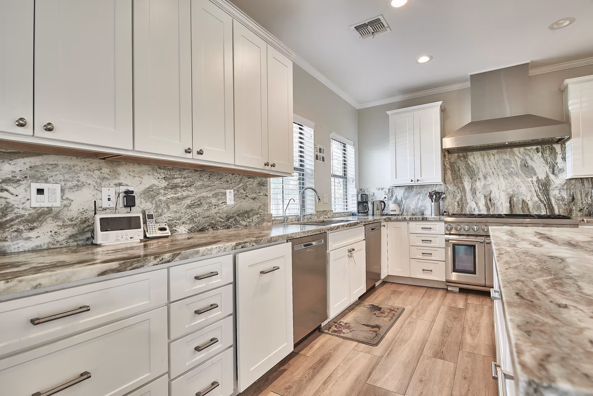 Modern kitchen with white cabinets, marble countertops and backsplash, stainless steel appliances including a stove with hood, dishwasher, and sink under two windows. The floor is wood and there are small kitchen appliances on the counter.