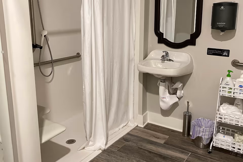 Accessible bathroom showing a walk-in shower with curtain and grab bar, a wall-mounted sink and mirror, and a shelving cart with toiletries.