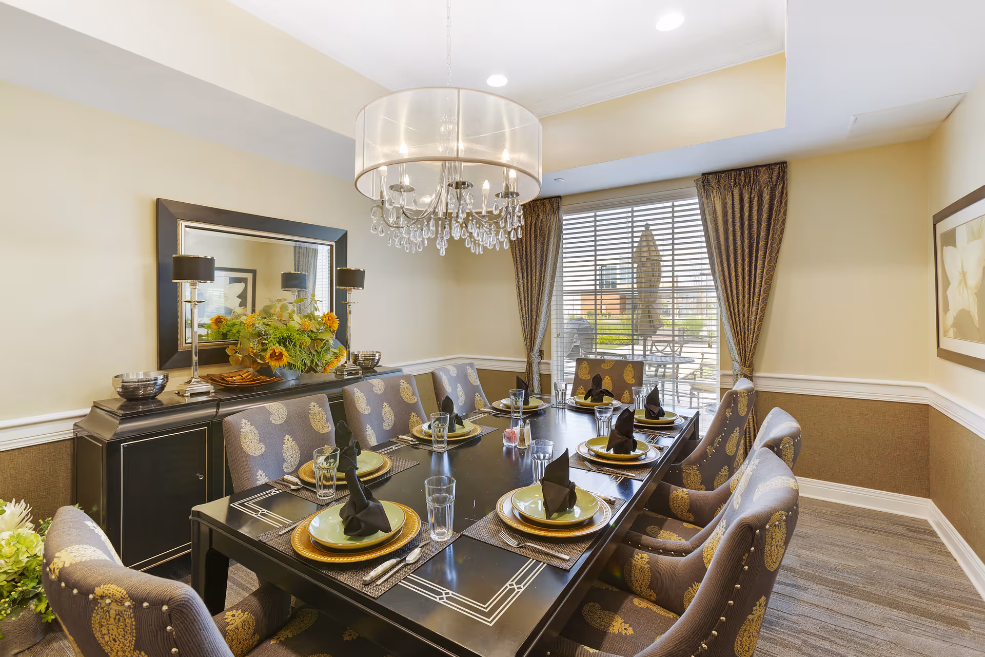 A well-lit dining room with a long black dining table set for eight people. Each place setting includes a green plate on a gold charger, a glass, silverware, and a black folded napkin. The chairs are upholstered with a patterned fabric in shades of gray and gold. A large mirror and two black lamps with silver bases are on a sideboard against the wall. A chandelier with a translucent drum shade and hanging crystals hangs above the table. Large windows with brown curtains reveal an outdoor patio with tables and umbrellas.