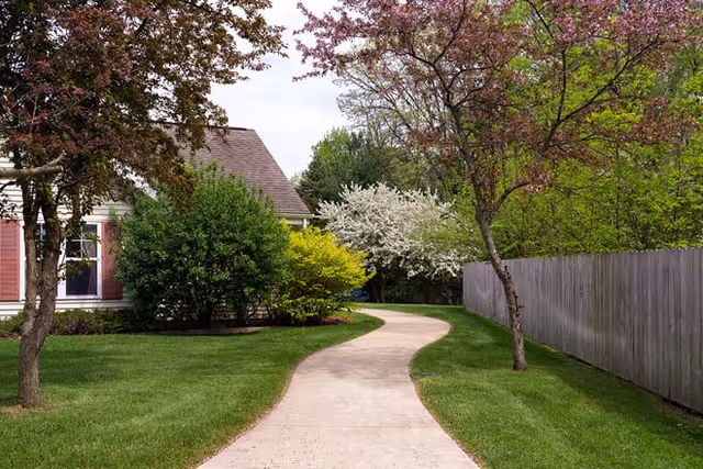 A curved concrete pathway leading through a green lawn with trees and bushes on either side. A house with red shutters is partially visible on the left, and a wooden fence runs along the right side of the path. Trees with pink and white blossoms are present, indicating a springtime setting.